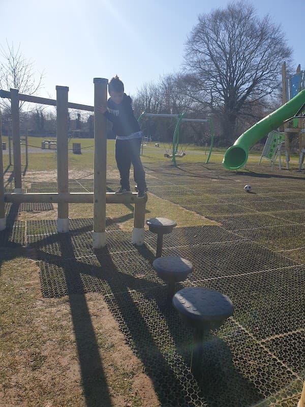 A child climbing on wooden playground equipment with a slide in the background, set in a grassy area under clear blue skies.