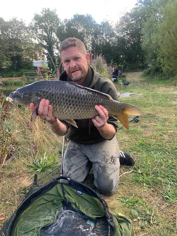 Man holding a large fish by Brookfoot Lake, surrounded by greenery and fishing gear in the background.