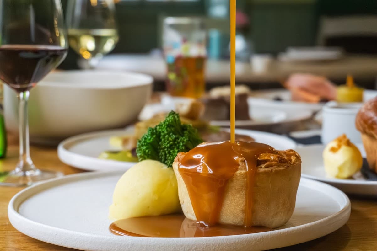 A plate with a meat pie drizzled in gravy, alongside mashed potatoes and broccoli, with wine glasses in the background.