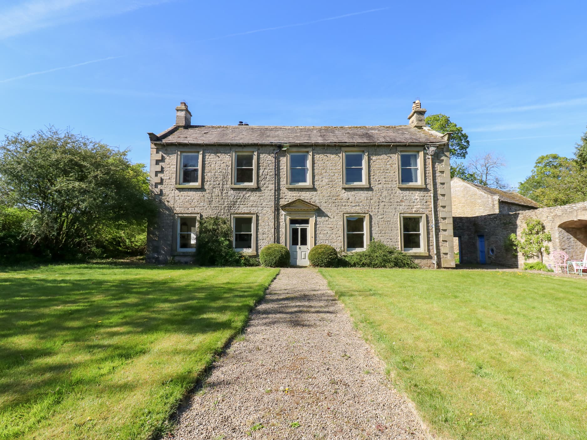 Two-story stone manor with chimneys, surrounded by lush green lawn, gravel path leading to entrance, and blue sky.