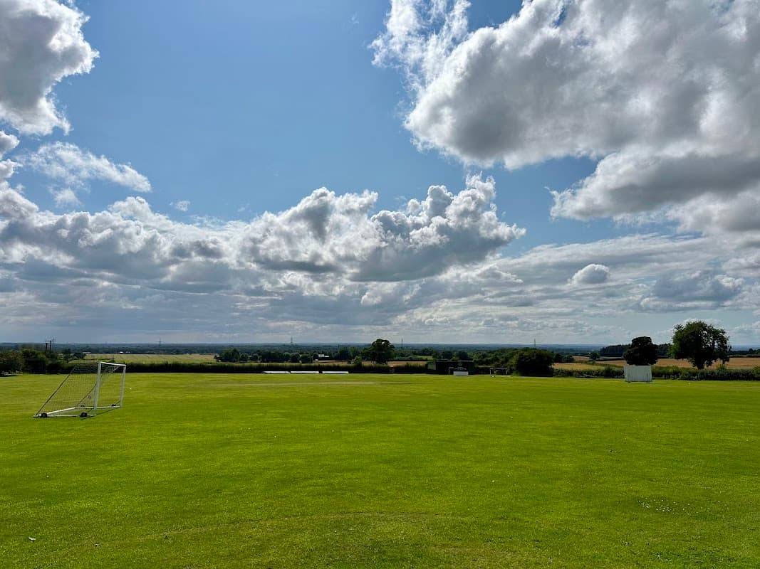 Green sports field with a goalpost, under a blue sky filled with clouds, overlooking a rural landscape.