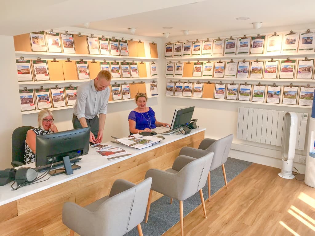 Bright office space with three staff members at desks, showcasing property listings on clipboards on the wall.