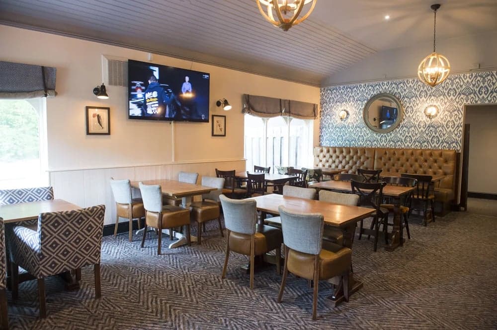 Cozy restaurant interior with wooden tables, chairs, patterned wallpaper, and a large TV on the wall.