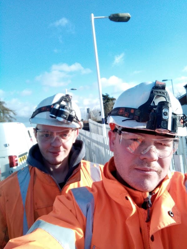 Two workers in orange safety jackets and helmets with headlamps, standing near a bus stop under a clear blue sky.