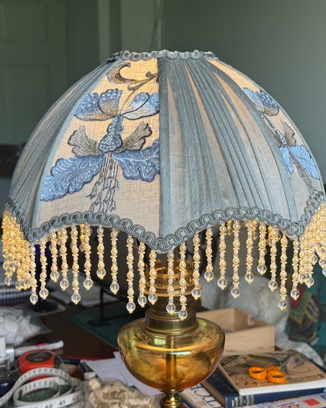 A decorative lamp with a blue floral shade and beaded fringe, resting on a brass base, in a bright interior setting.