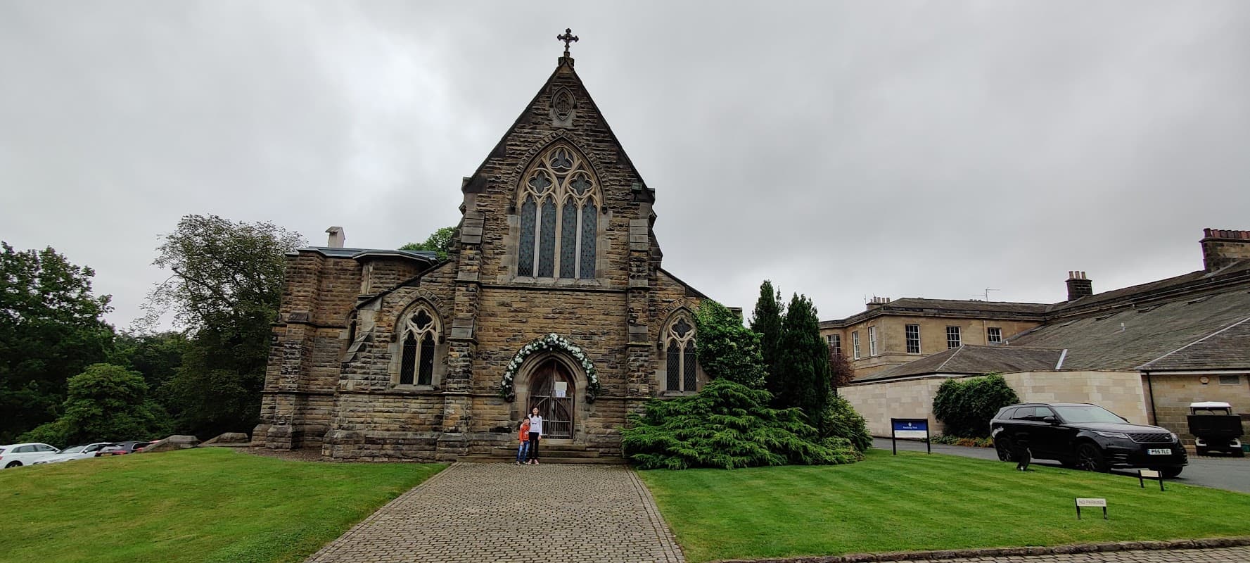 Victorian-style church with stone faรงade, arched windows, and a cross atop, surrounded by greenery and a gravel path.