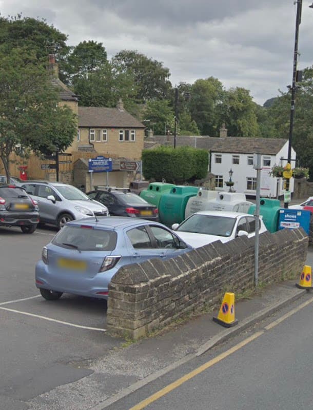 Honley Westgate Car Park with parked cars, stone wall, and nearby buildings under a cloudy sky.