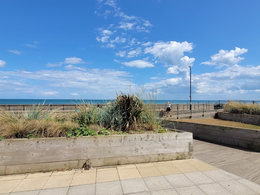Hornsea Hub with wooden planters, coastal views, and a clear blue sky, featuring a person walking along the promenade.