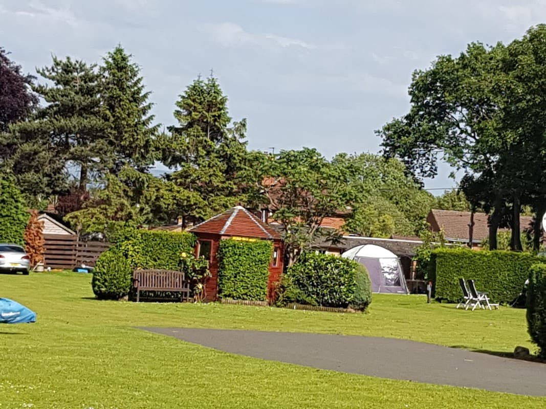 Lush green lawn with a gazebo, benches, and tents, surrounded by trees and a clear blue sky at Pembroke Caravan Park.