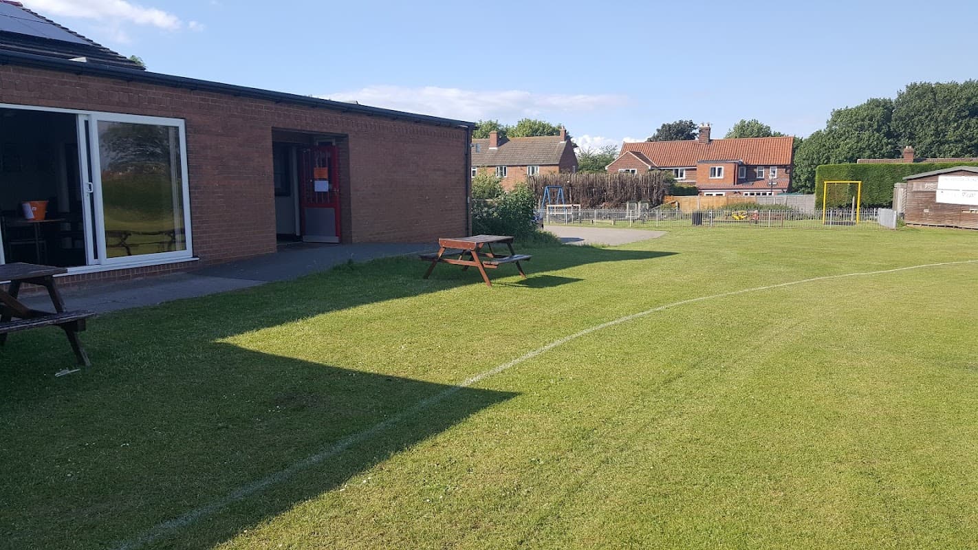 Long Marston Village Hall with grassy area, picnic tables, and neighboring houses under a clear blue sky.