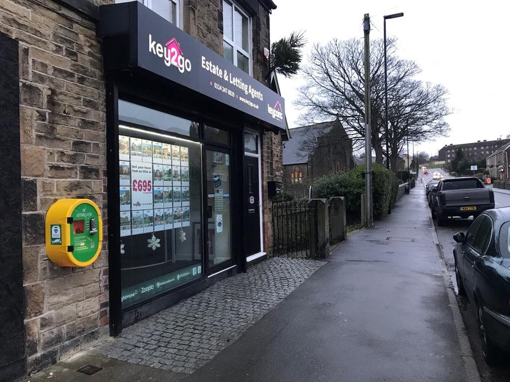 Defibrillator mounted on a wall beside an estate agency, with a wet pavement and trees lining the street in Mosborough.