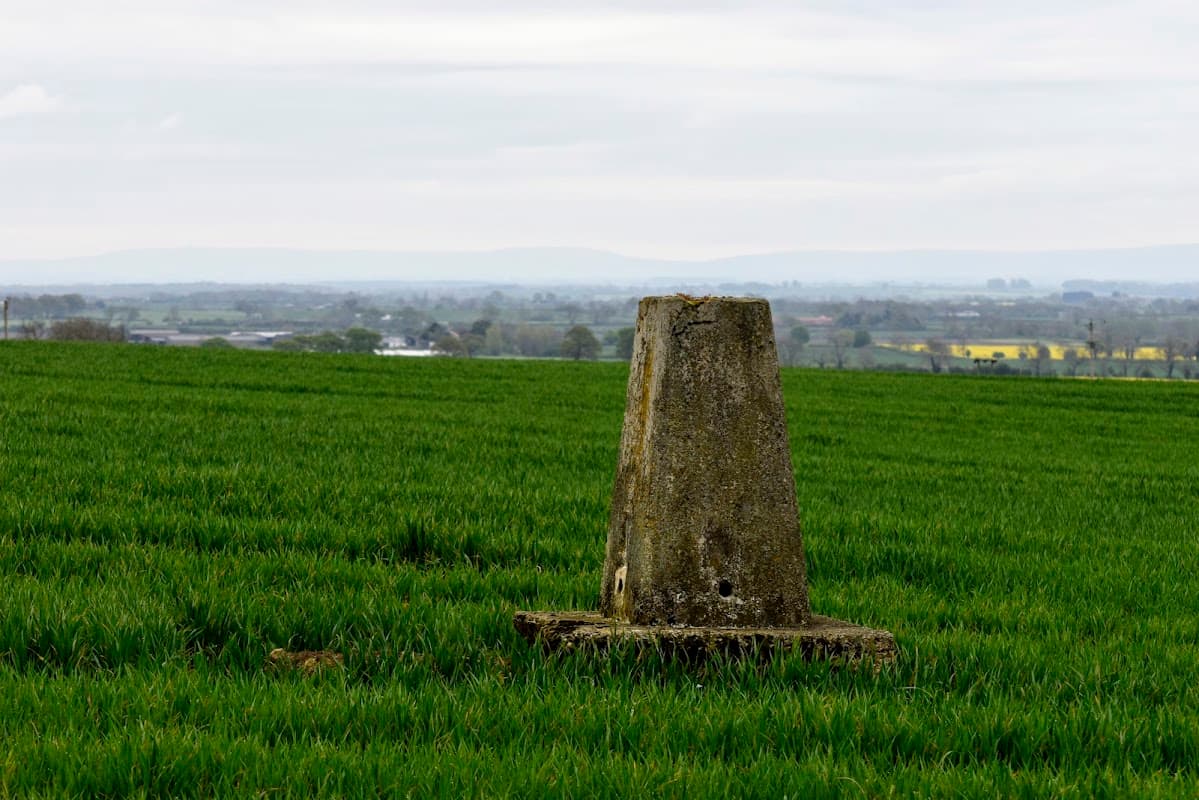 Trig point in a green field with distant hills and a cloudy sky in Moulton, North Yorkshire.