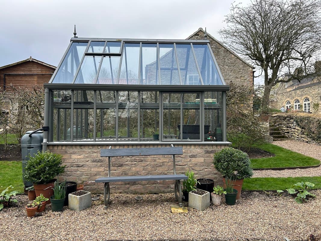 Glass greenhouse with stone base, surrounded by potted plants and a bench, set in a garden with stone buildings.