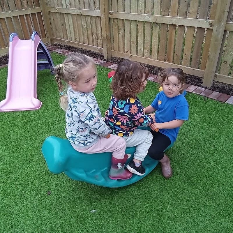 Three children sit on a green rocking toy in a playground, with a pink slide and wooden fence in the background.
