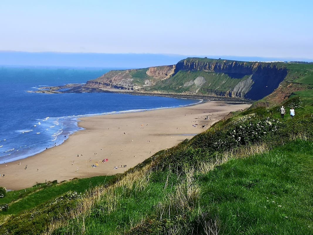 Cayton Bay with sandy beach, cliffs, and green hills under a clear blue sky, featuring people enjoying the shore.