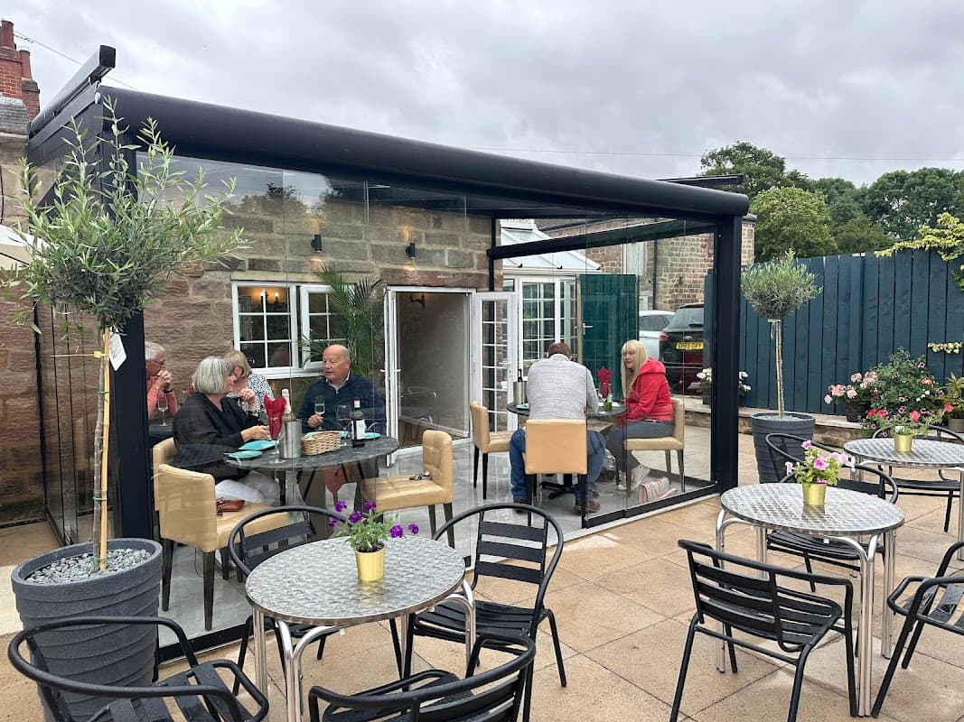 Glass-walled bistro with outdoor seating, guests enjoying drinks, and potted plants in a cozy setting.