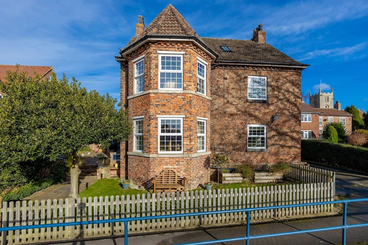 Charming brick boutique B&B with large windows, a garden bench, and a picket fence under a clear blue sky.