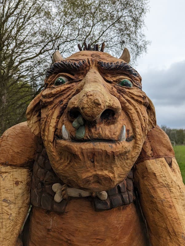 Wooden sculpture of a troll with exaggerated features, set against a backdrop of trees and cloudy skies in Oxley Park.