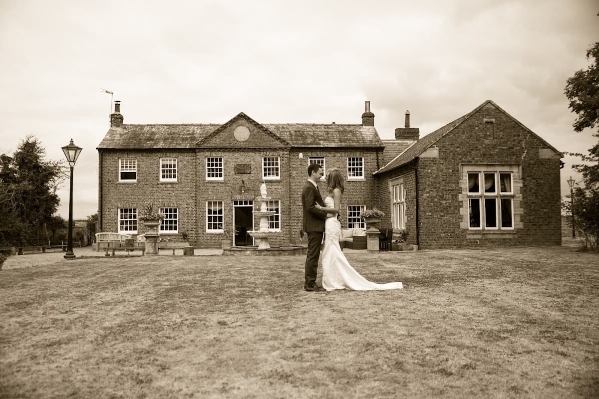 Historic brick building with large windows, a couple in formal attire stands on the lawn, surrounded by greenery.