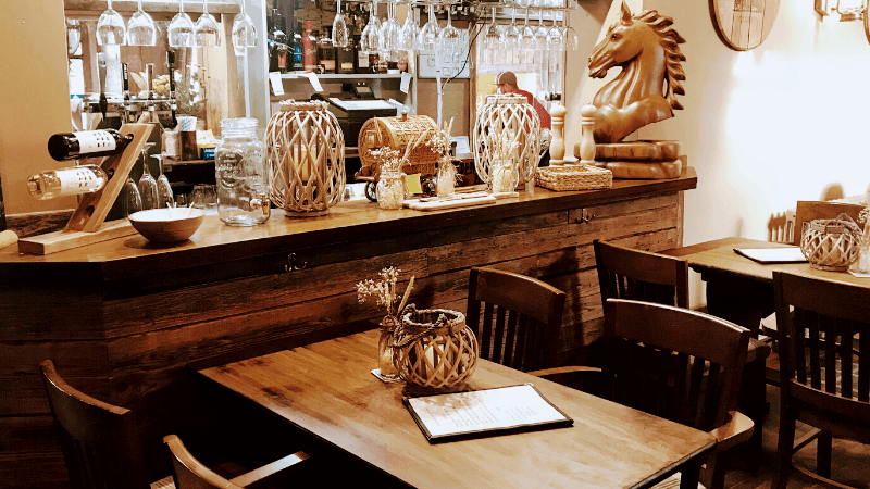 Rustic restaurant interior featuring wooden tables, a bar with decorative jars, and a wooden horse sculpture.