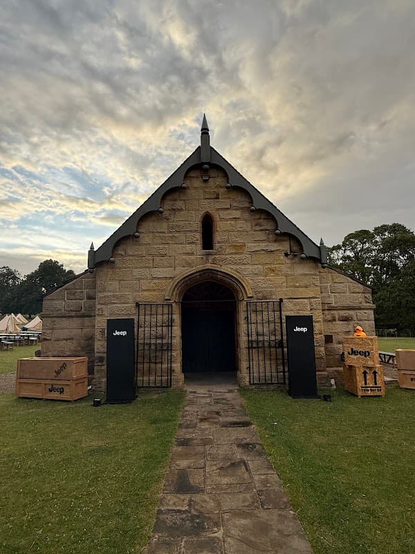 Historic stone building with a pointed roof, flanked by black columns and wooden crates on the lawn under a cloudy sky.