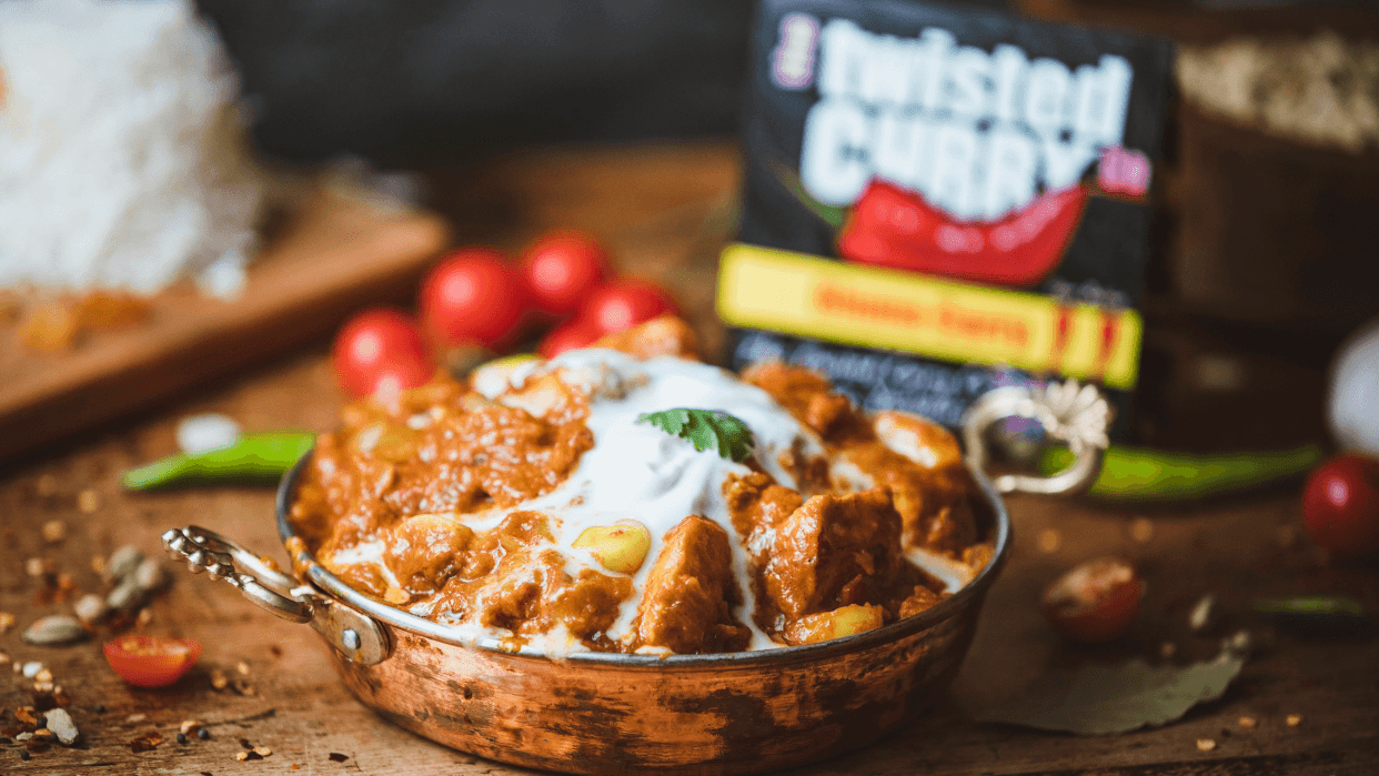 A copper bowl filled with rich curry topped with yogurt and herbs, surrounded by fresh tomatoes and spices.