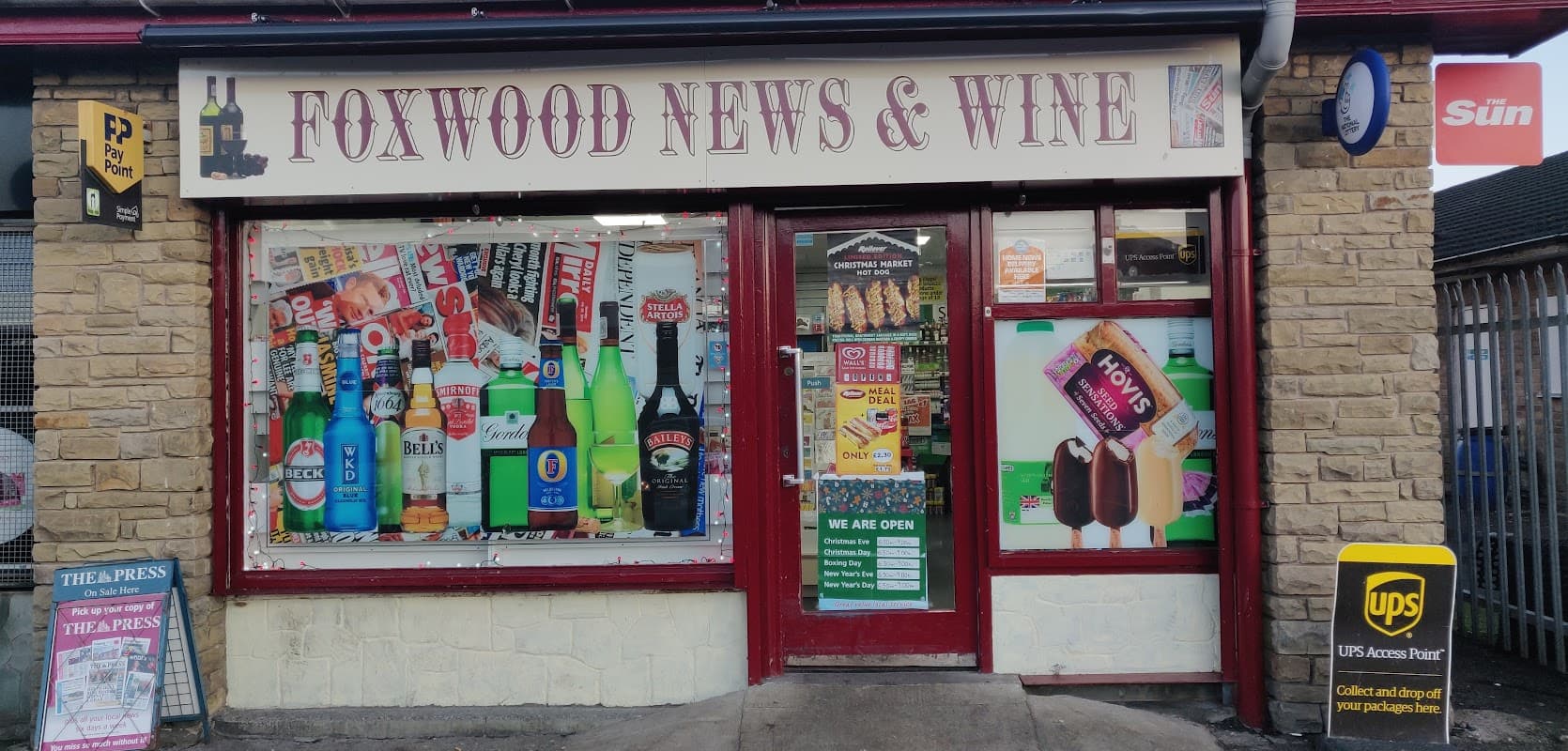 Foxwood Newsagents & Wine shopfront with colorful advertisements, drinks, and a sign stating "WE ARE OPEN."