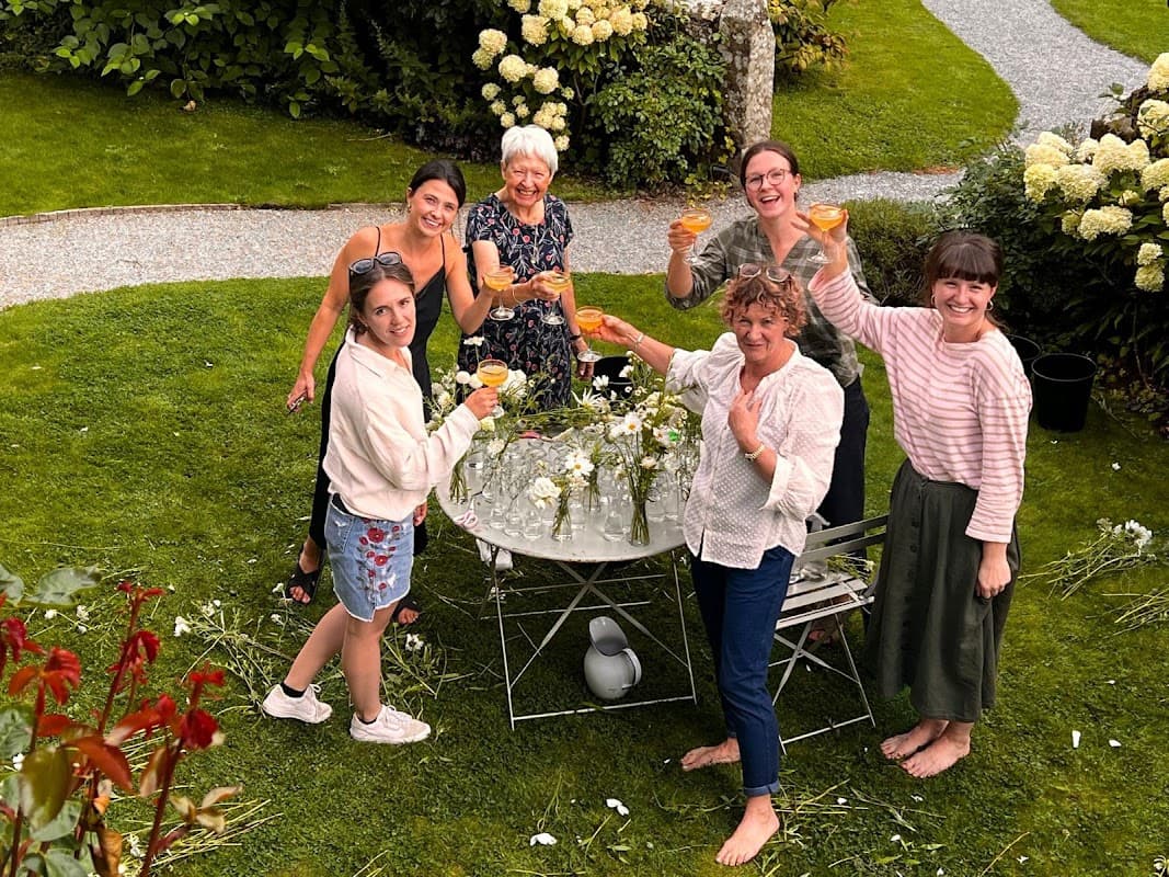 A group of six women celebrating with drinks in a garden, surrounded by flowers and greenery.