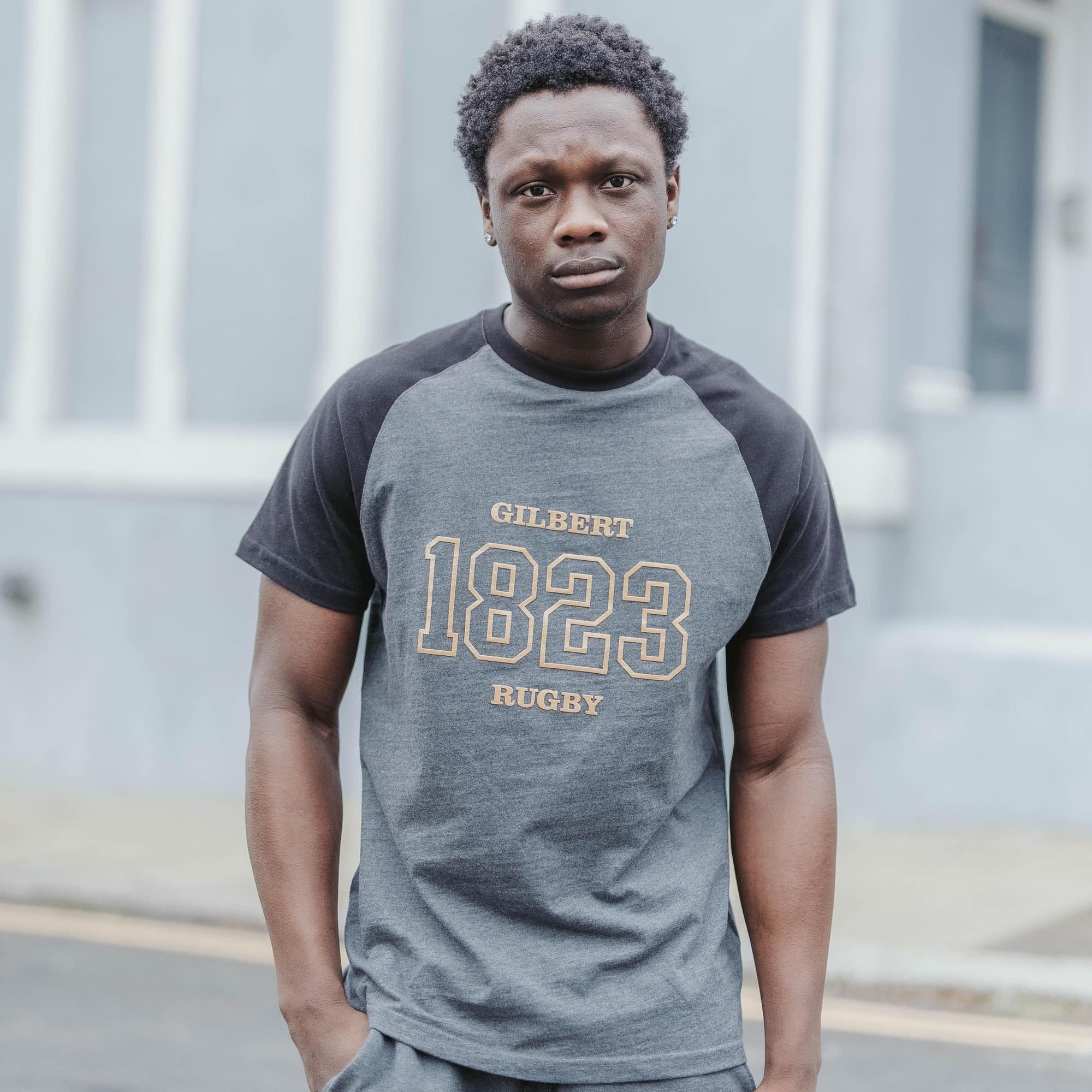 Gray and black short-sleeve t-shirt featuring "GILBERT" and "1823 RUGBY" printed in gold on the front, made from soft cotton material, suitable for casual wear.