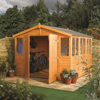 A brown wooden workshop measuring 9ft x 15ft, featuring double doors, large windows, and a sloped roof, surrounded by greenery and potted plants.