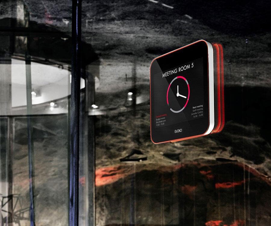 Evoko Liso is a sleek black and red digital meeting room display with a circular clock interface, mounted on a glass wall in a modern setting.