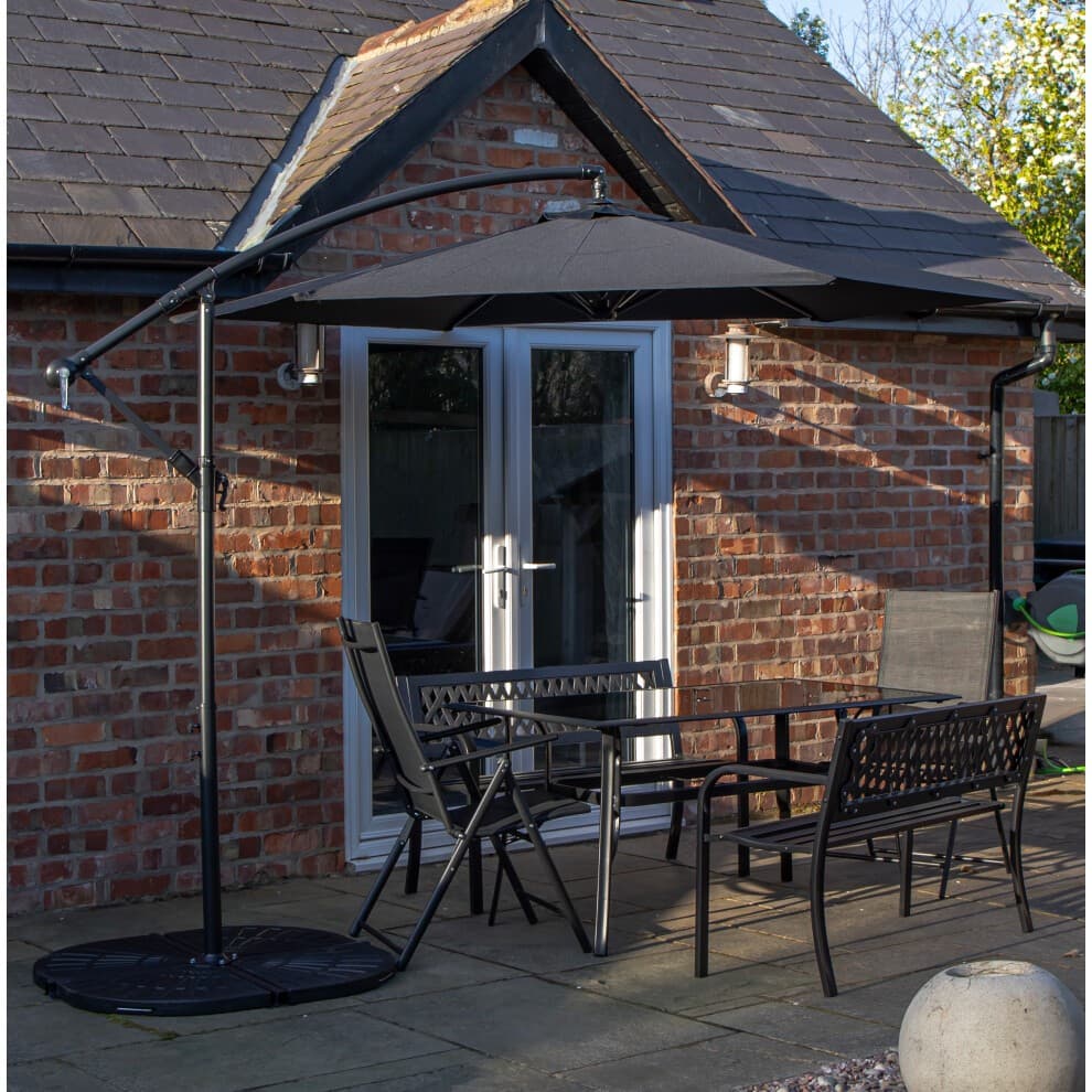 Rectangular glass top table with black metal benches and chairs, accompanied by a black banana parasol and base, set against a brick wall and patio.