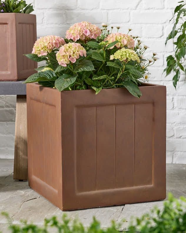 Square brown planter made from durable material, featuring a textured design and filled with pink and green hydrangeas, set against a white brick wall.