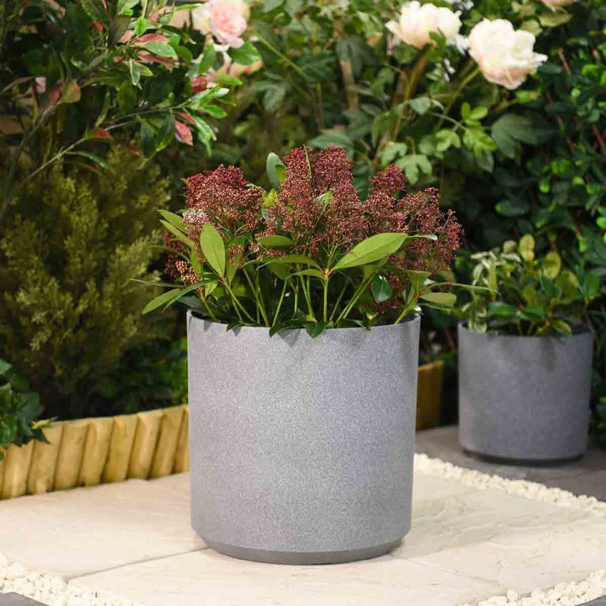 Grey 26cm cylindrical planter made of durable material, featuring a textured finish and filled with lush green foliage and red flowers, set against a garden backdrop.