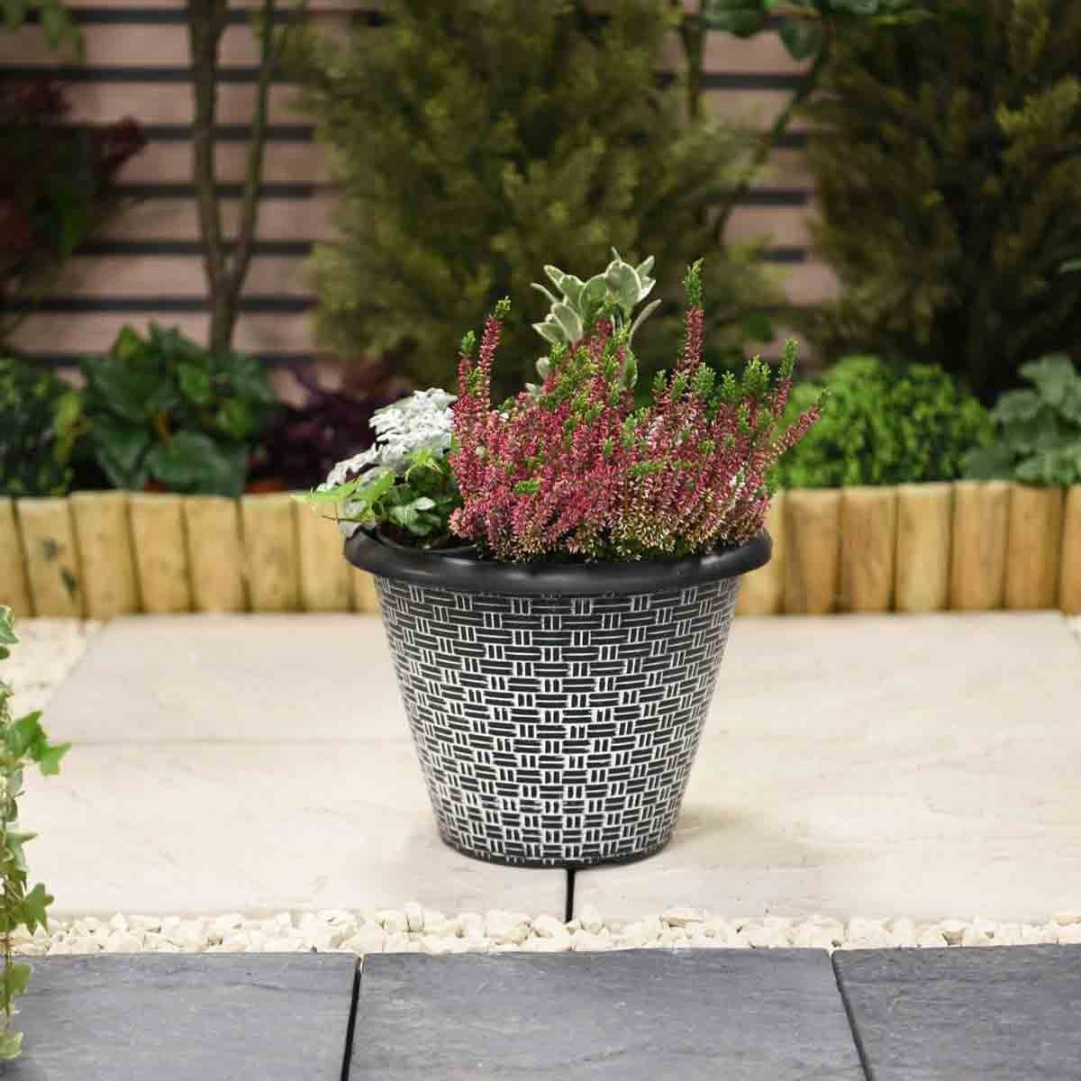 Grey 30.5cm planter with a woven effect design, featuring a variety of colorful plants, set against a landscaped garden background.