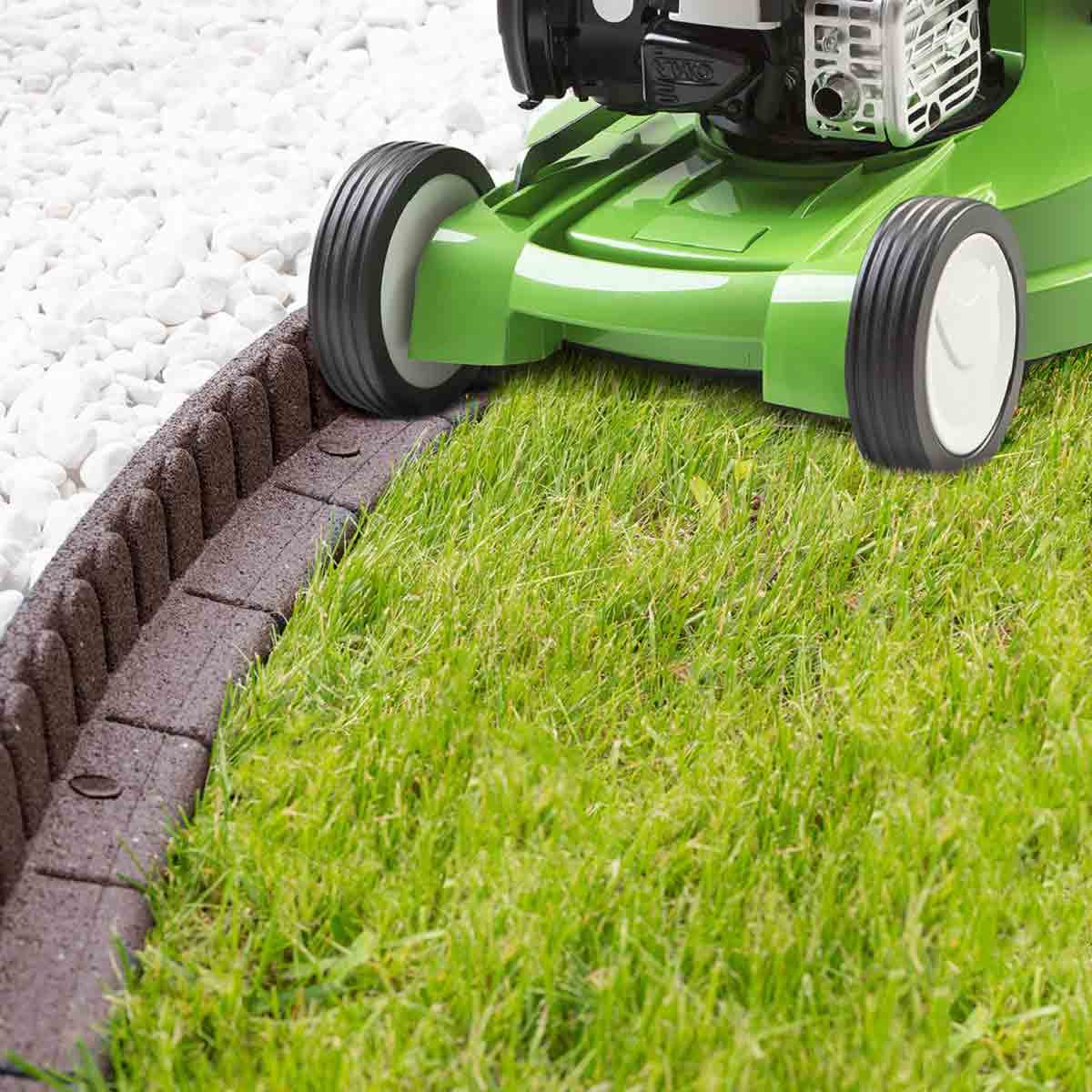 Green lawnmower next to brown, textured border edging made of durable material, outlining a neat grass area, with white gravel in the background.