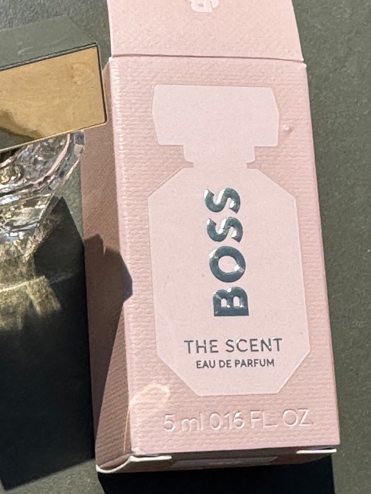 Pink box featuring "BOSS THE SCENT" in silver lettering, with a clear glass bottle and gold cap, labeled 5ml Eau De Parfum.