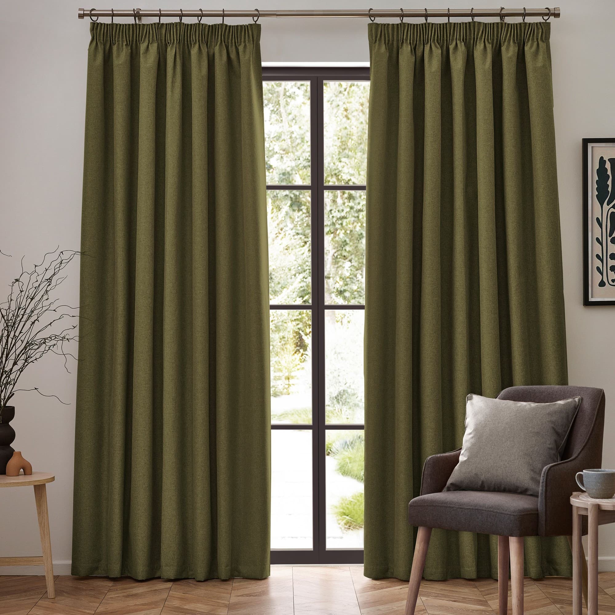 Olive green blackout pencil pleat curtain pair, measuring 168cm width by 137cm drop, hanging from a rod in a well-lit room with a plant and a chair.