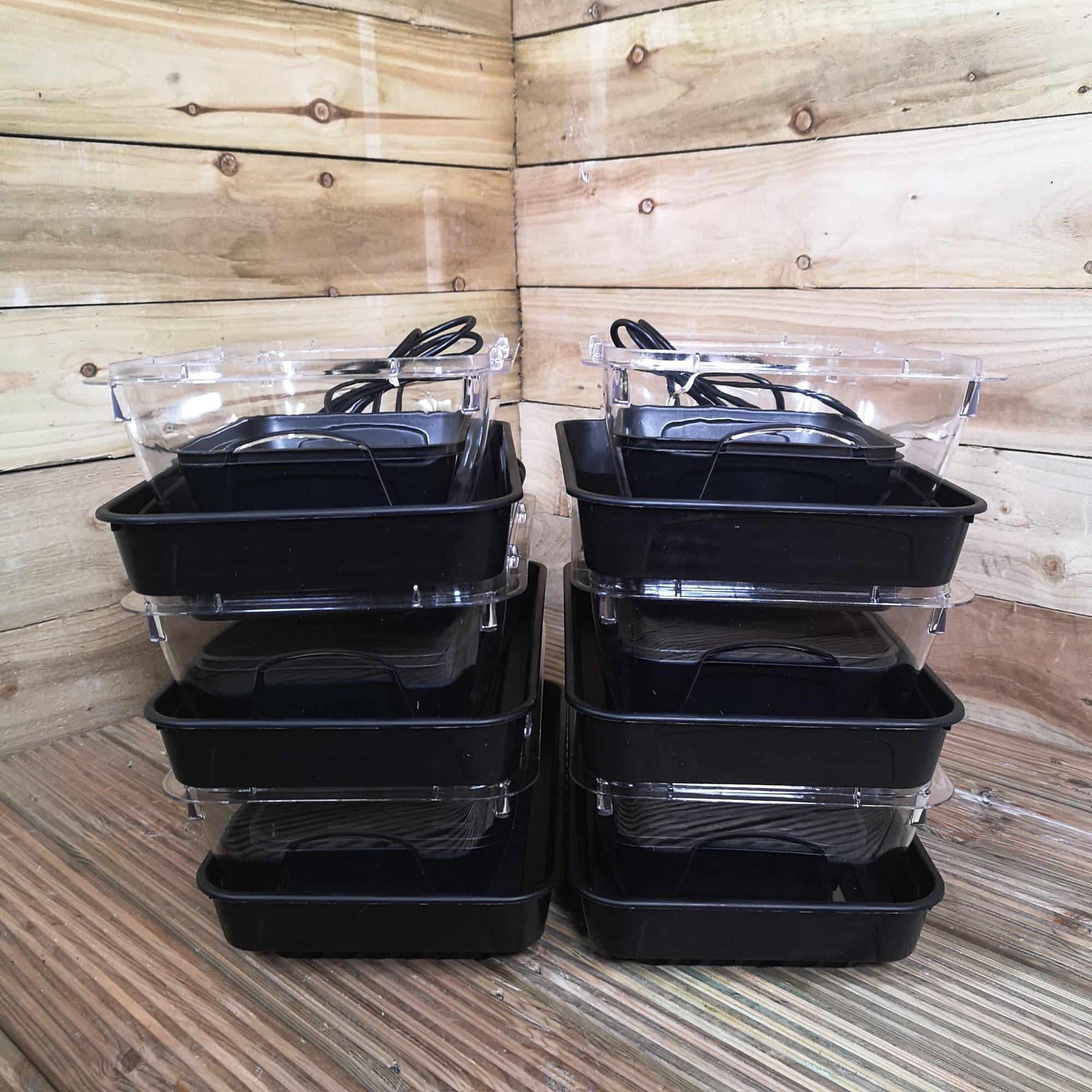 Two stackable black plastic seed starter trays with transparent lids, featuring heating elements, designed for indoor seedling propagation, arranged neatly on a wooden surface.