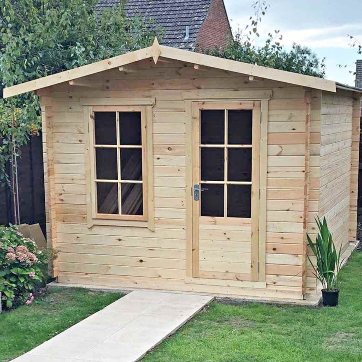 Light wood log cabin summerhouse with a sloped roof, double front doors, and two windows, set in a garden with greenery and a stone pathway.