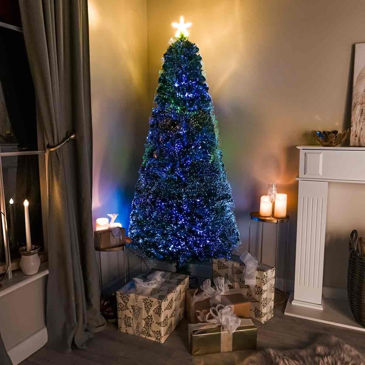 5ft green fibre optic Christmas tree with pin prick design, illuminated with multicolored lights, placed near wrapped gifts and candles in a cozy room setting.