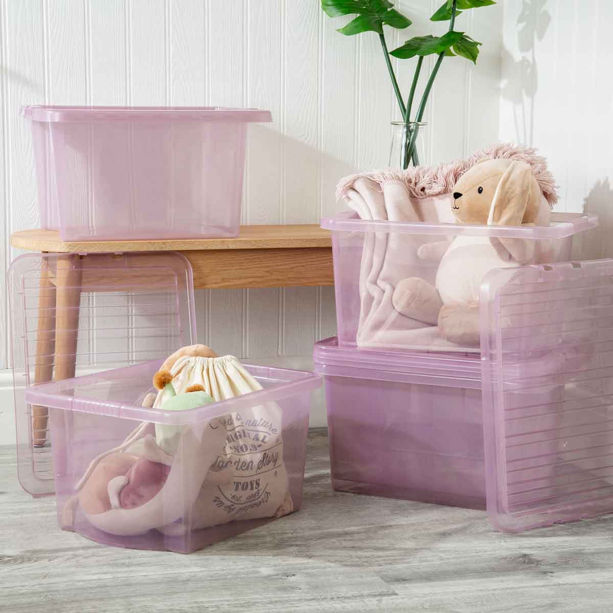 Set of five pink transparent plastic storage boxes with lids, featuring a ribbed design, stacked and filled with soft toys and blankets, placed beside a wooden table and a plant.