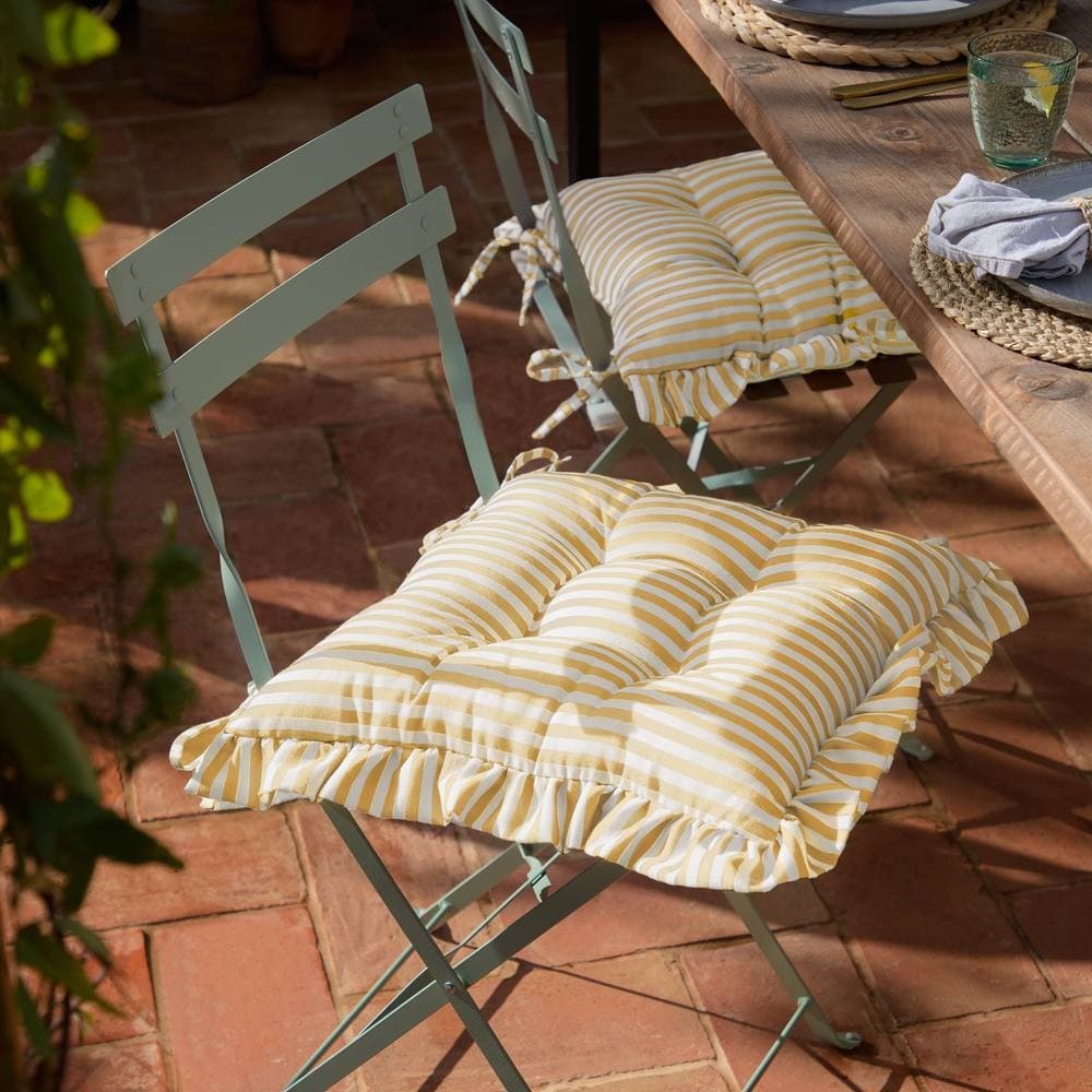 Yellow striped ruffle seat pads with tufted design, placed on light blue metal chairs, enhancing a rustic outdoor dining setting.