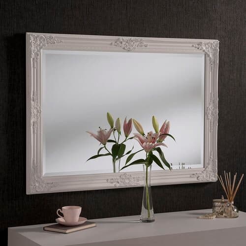 Decorative white mirror with an ornate frame, measuring 104 x 74 cm, reflecting a vase of pink lilies and a cup on a sleek surface against a dark wall.