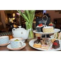 Afternoon tea set with teapot, floral cups, tiered tray of cakes, sandwiches, and a vase of tulips.