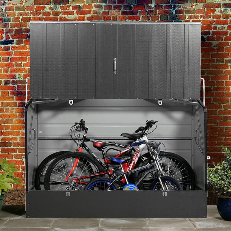 Secure garden bike storage in anthracite grey, made of metal, featuring a hinged top, two bicycles inside, and a brick wall backdrop.