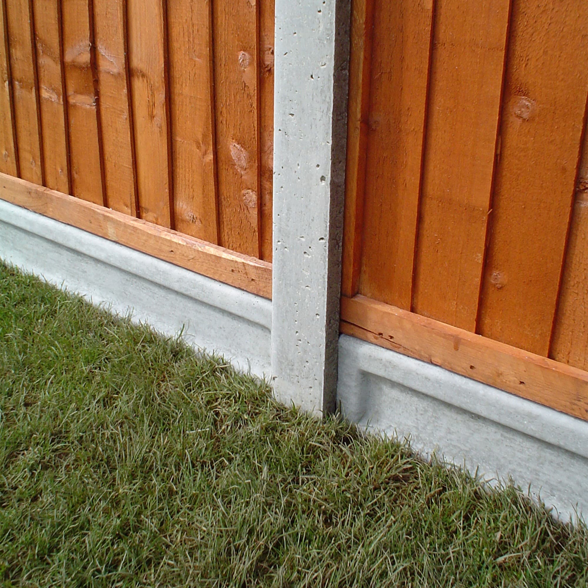 Lightweight concrete gravel board in grey, measuring 6' x 6", positioned at the base of a wooden fence with a warm orange finish, set against green grass.