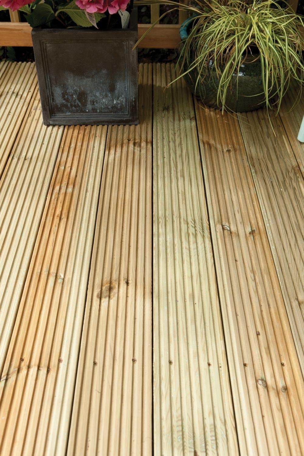 Light-colored, treated softwood deck boards arranged on a wooden surface, showcasing a smooth finish and visible grain, with a potted plant and decorative item nearby.
