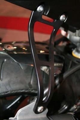Black metal motorcycle exhaust hanger with a sleek, curved design and multiple cutouts, featuring silver mounting screws, attached near a tire.