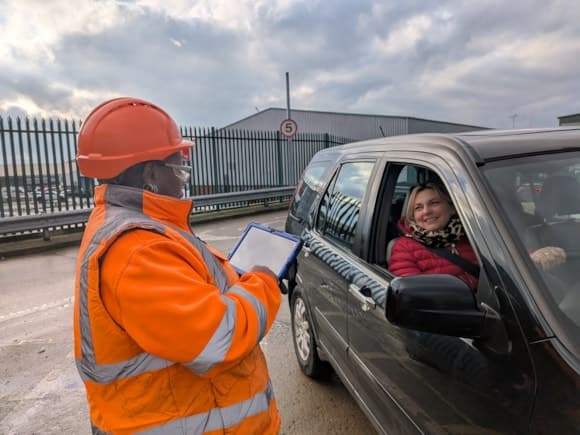 A worker in an orange safety vest and hard hat assists a smiling woman in a car at Hazel Court HWRC under a cloudy sky.